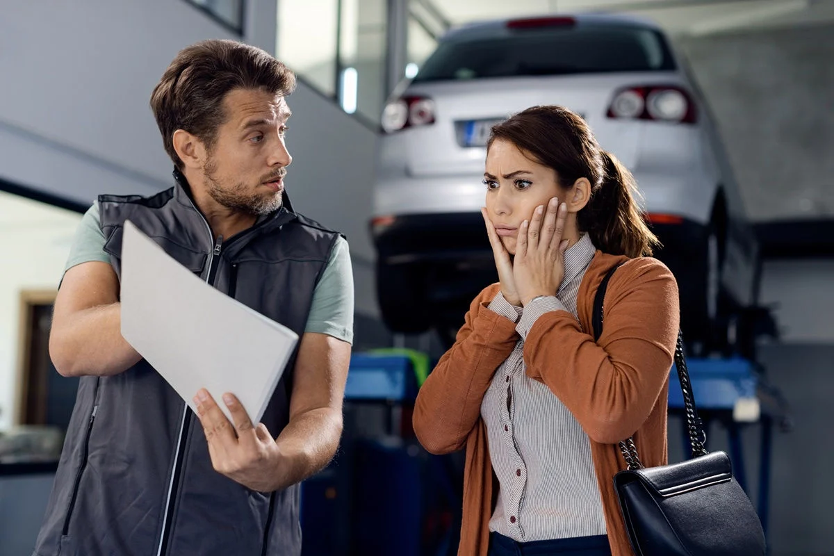 Photo of a car owner with a surprised look on her face as she looks at the repair order shown to her by an auto mechanic